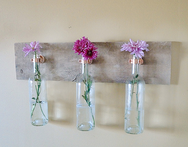 3 garrafa de vinho transparente com flores e penduradas numa madeira na parede